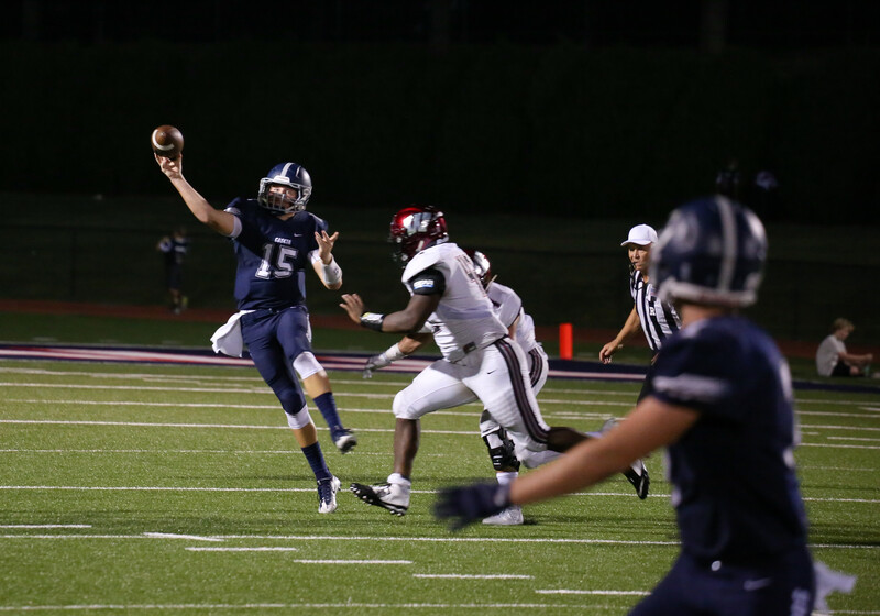High school football player in dark blue jersey throwing a pass while evading a defender in white during a night game.