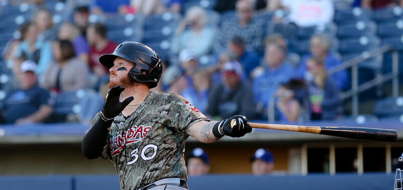 Baseball player in camo uniform mid-swing during game with crowded stadium behind.