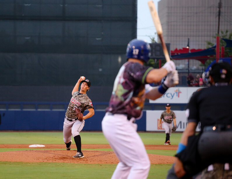 Baseball pitcher mid-delivery on the mound during a game, with batter and catcher in the background.