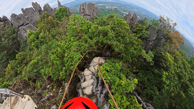 First-person view from a climber ascending a steep rocky peak covered in dense green vegetation.