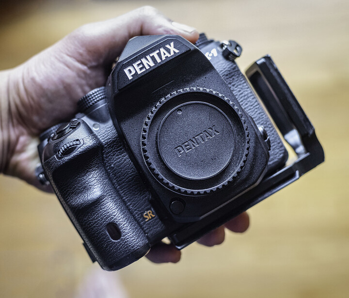Pentax DSLR camera body held in someone's hand against a blurred outdoor background.