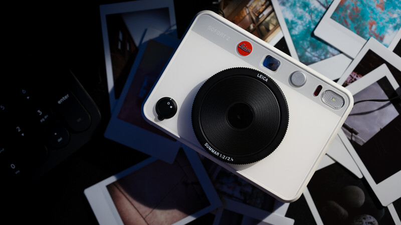 White hybrid instant and digital camera surrounded by printed photos on dark background.