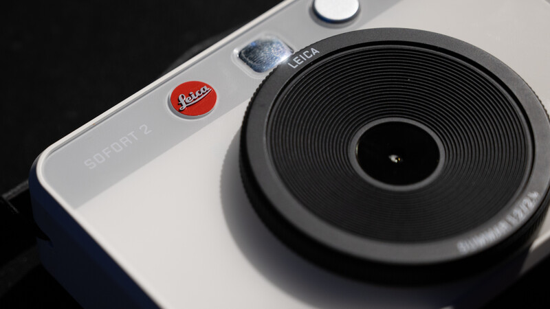 Close-up of Leica Sofort 2 instant camera showing red shutter button, Leica branding, and large black lens against white and dark body.