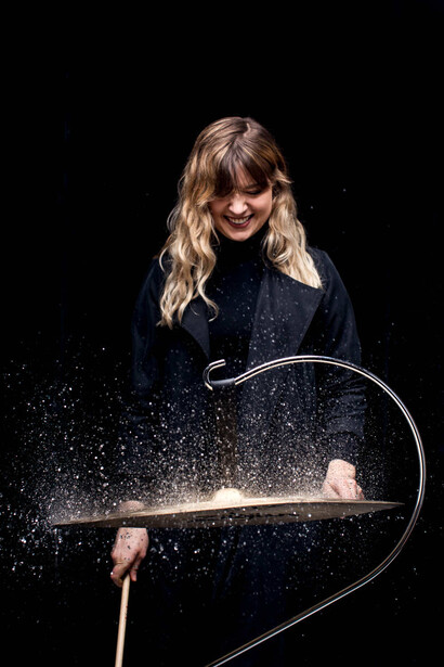 Person with blonde hair and red lipstick playing drums against a black background with dramatic lighting and water spray effects.