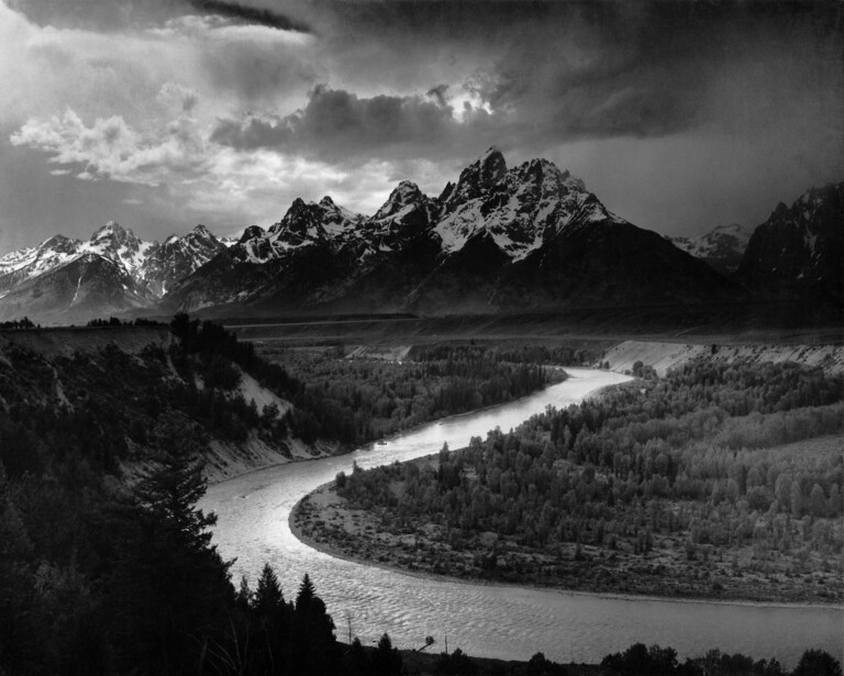 Dramatic black and white landscape featuring snow-capped mountains, a winding river, and dense forest under stormy skies.