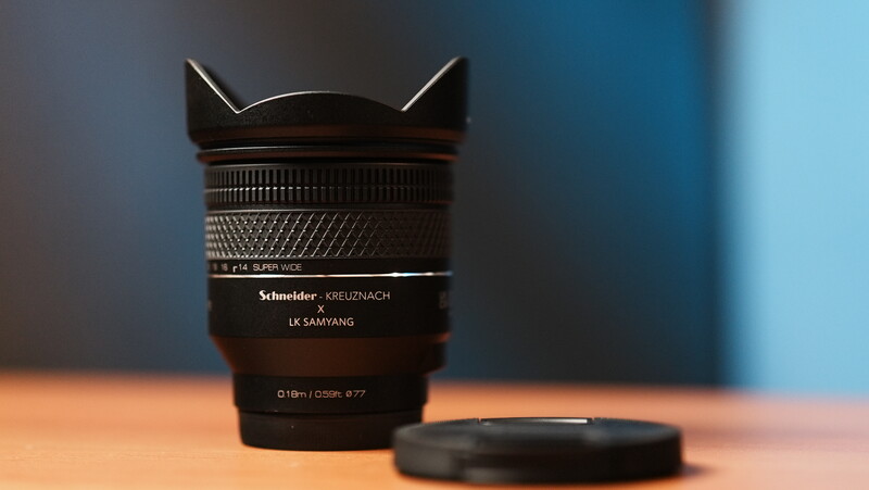 Schneider Kreuznach LK Samyang lens with lens cap on wooden surface against blue background.