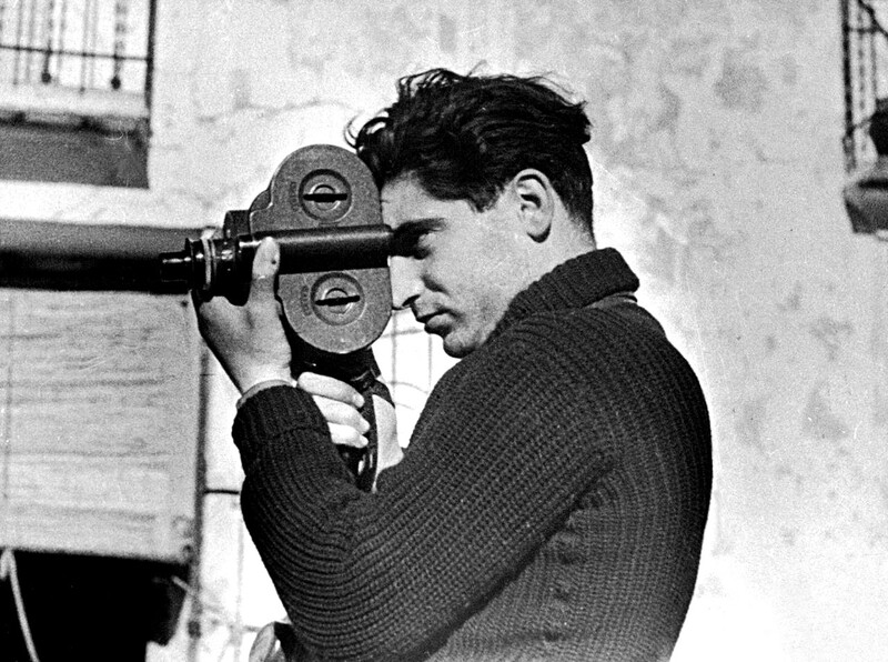 Man in profile holding a film camera to his eye, positioned against a weathered interior wall.