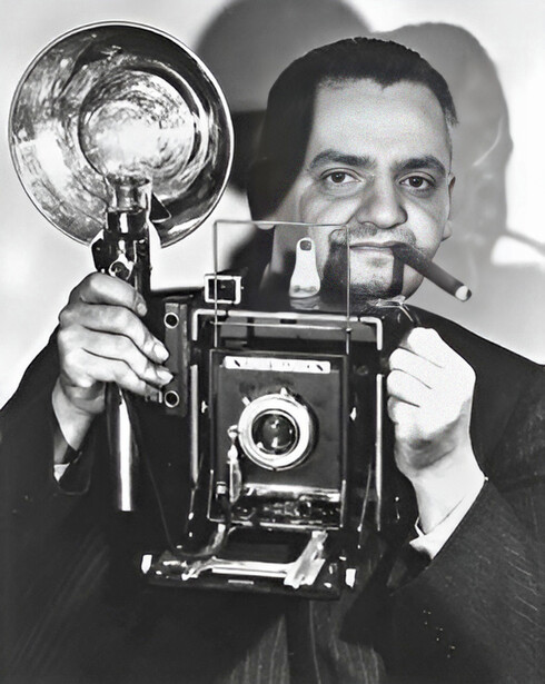 Photographer holding a large format camera and flash reflector, posing for a black and white portrait.