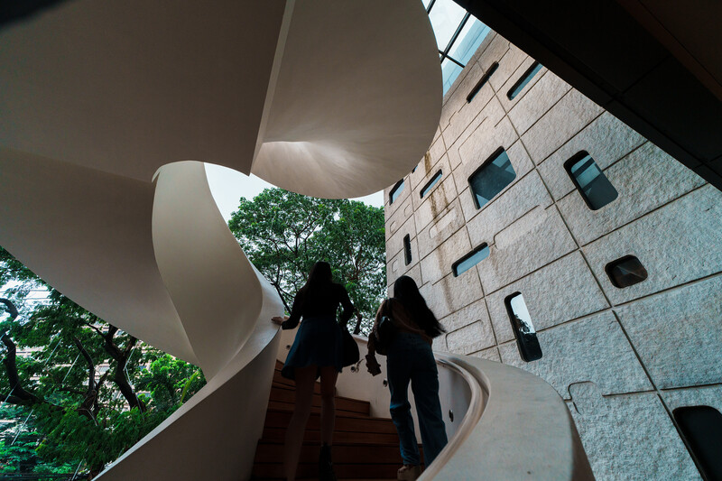 Two silhouetted figures ascending a curved concrete staircase beneath a sculptural overhang, with a modern stone building facade visible above.