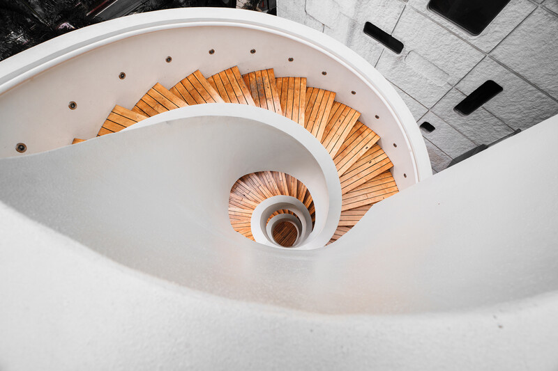 Spiral staircase with white walls and orange-brown wooden steps viewed from above, creating concentric circular pattern.