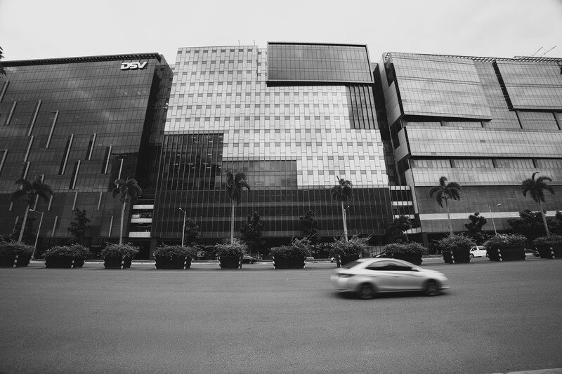 A blurred white car passes through a modern plaza lined with tall glass and steel buildings.