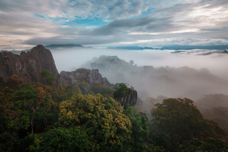 Dramatic mountain peaks rising above a thick sea of fog at dawn, with layered clouds and blue sky above.