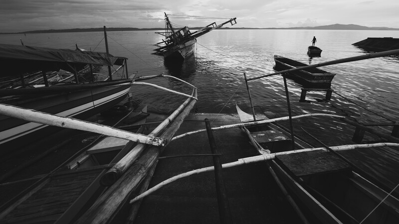 Moored fishing boats with wooden outriggers in calm water under overcast sky.