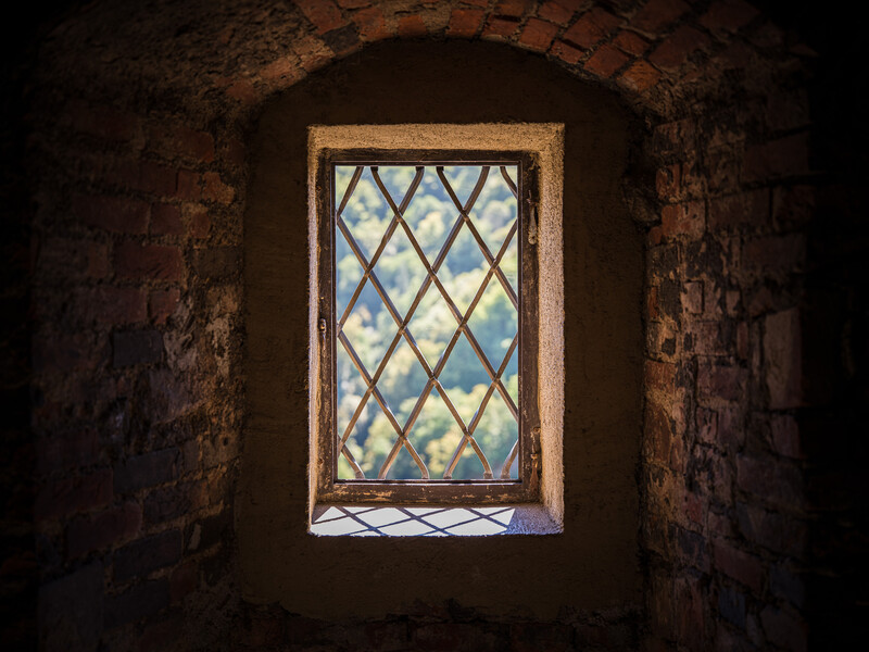 View through a diamond-patterned leaded window in an old brick archway, looking out to daylight and distant landscape.