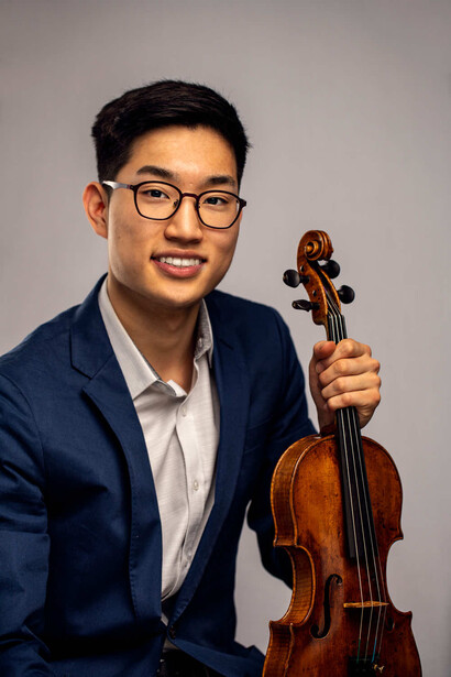 Young man in navy blazer and glasses holding a violin, photographed against a neutral gray background.