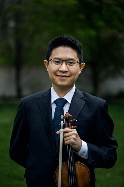 Professional musician holding a violin while wearing glasses and formal business attire outdoors.