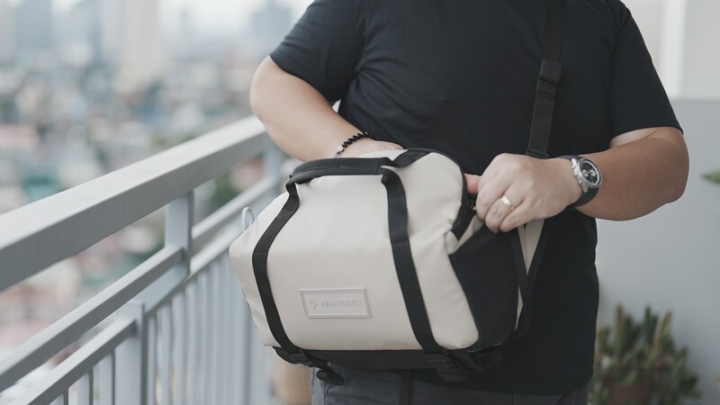 Man in black shirt holding white and black camera bag on high-rise balcony overlooking cityscape.