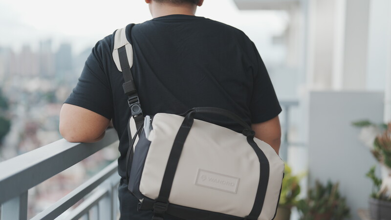 Person wearing a black t-shirt and two-tone camera bag standing on a high-rise balcony overlooking a cityscape.