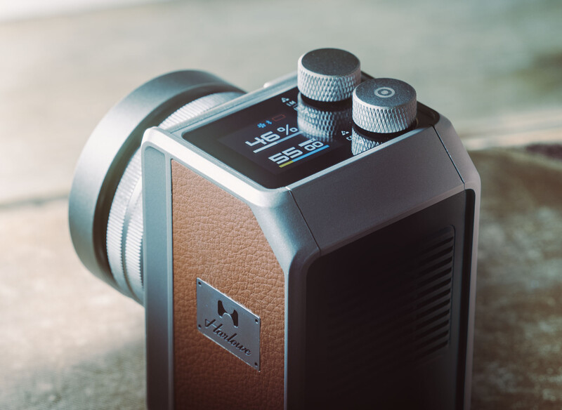 Compact portable audio device with brown leather panel, black casing, top-mounted screen, and two silver control knobs with shoulder strap.