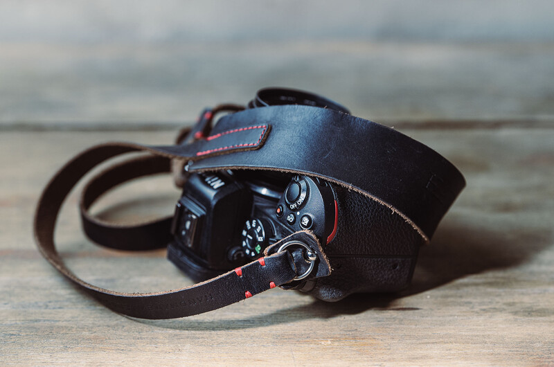 Dark leather camera strap and case with brown stitching detail on wooden surface.
