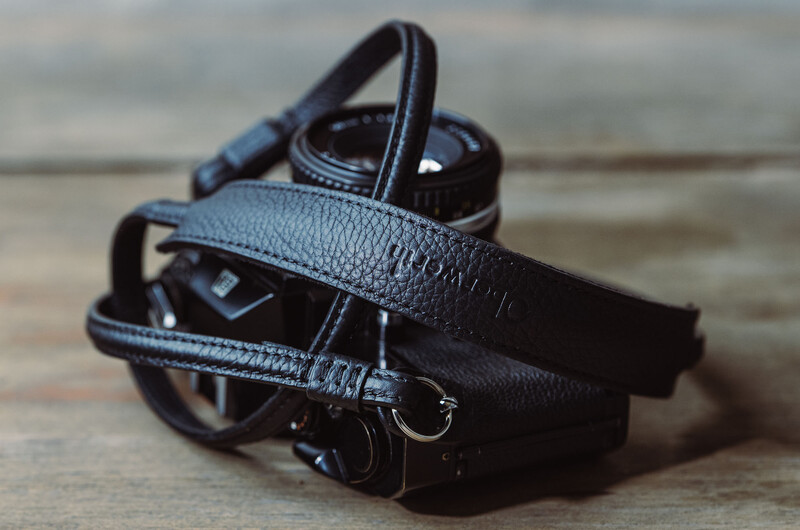 Black leather camera strap coiled on wooden surface with camera equipment in soft natural light.