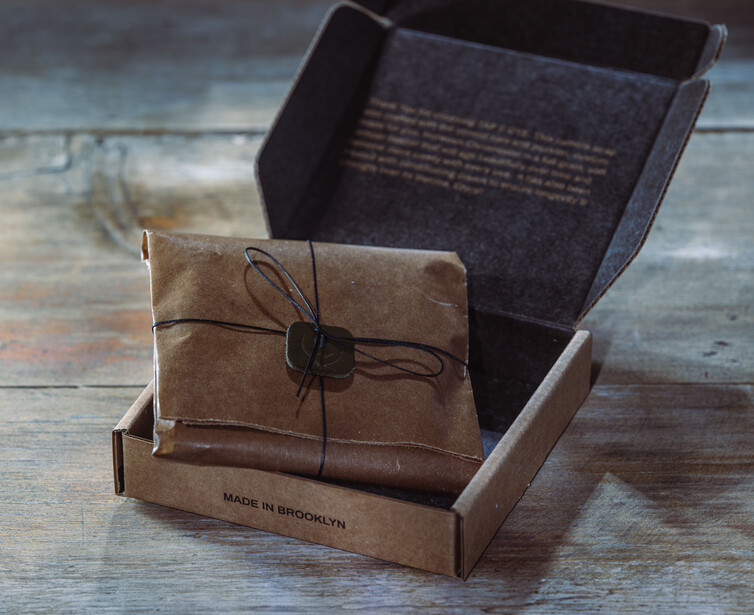 Kraft paper package with twine bow nested in open cardboard box on weathered wood surface.