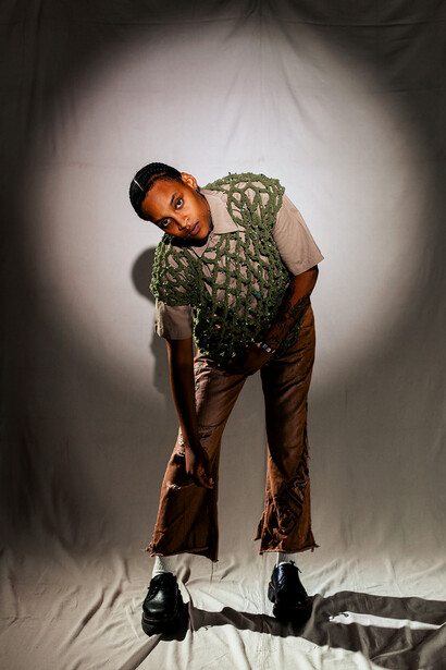 Person in patterned green shirt and brown pants posed against white seamless backdrop with studio lighting.