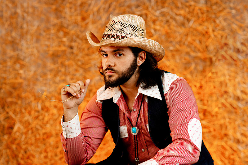 Man wearing cowboy hat and western-style clothing against textured orange backdrop.