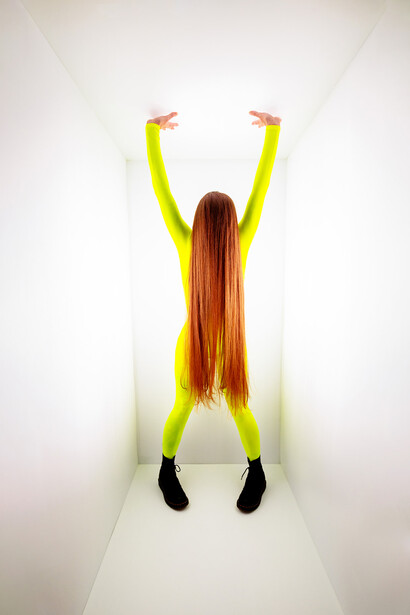Woman with long reddish-brown hair wearing neon yellow sleeves raising both arms overhead in a white corridor.