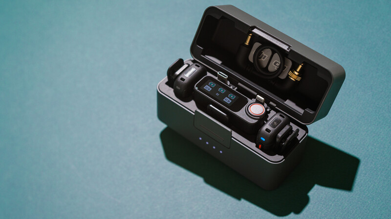 Camera gear laid out on turquoise surface including compact camera, wireless microphone, and storage case with accessories.