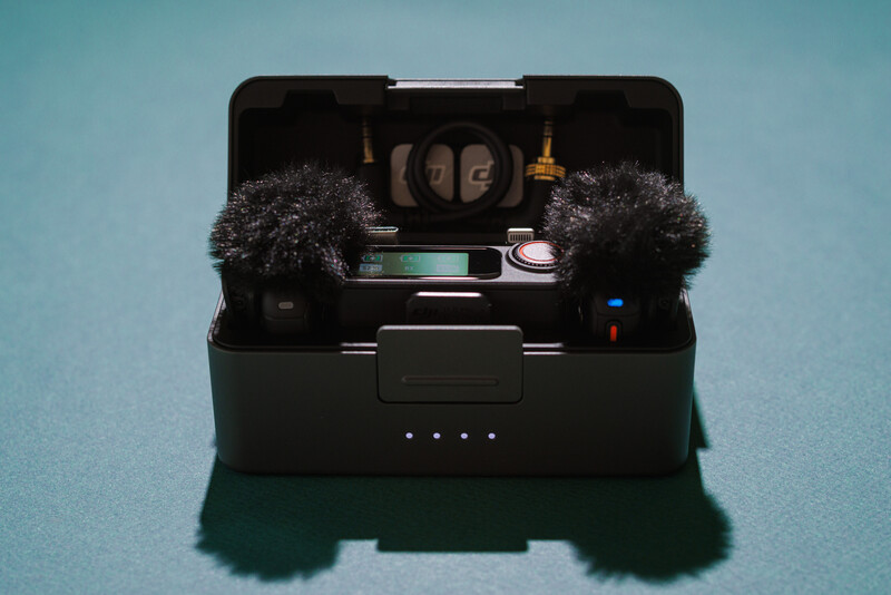 Open black field recording kit displaying microphones, windscreens, cables, and audio accessories on a blue background.