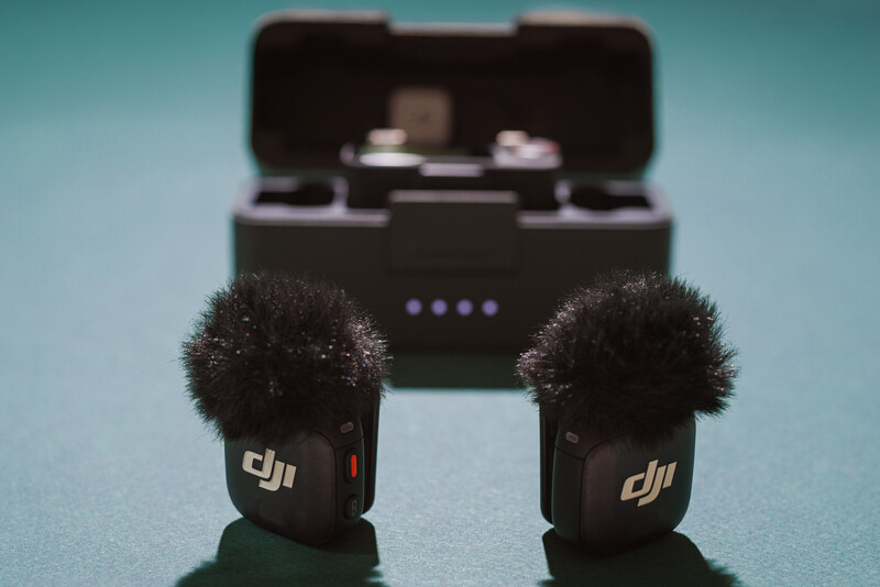 Two wireless microphones with fuzzy windscreens and their charging case against a blue background.