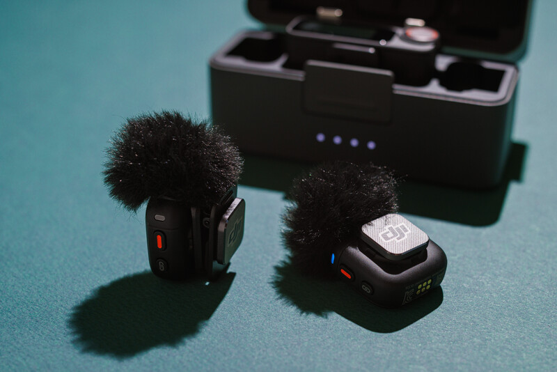 Two wireless microphone transmitters with fuzzy windscreens next to a black carrying case on a turquoise surface.