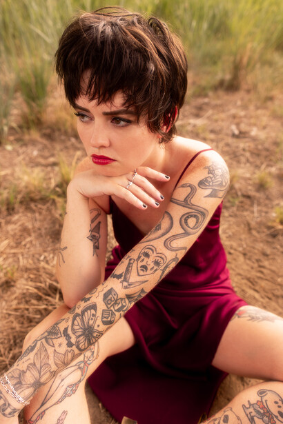 Woman with short dark hair and tattoo-covered arms posing thoughtfully in an outdoor field setting.