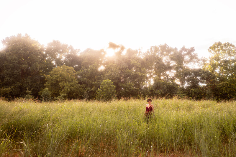 Solitary figure standing in a vast golden grass field at golden hour with tree line in background.