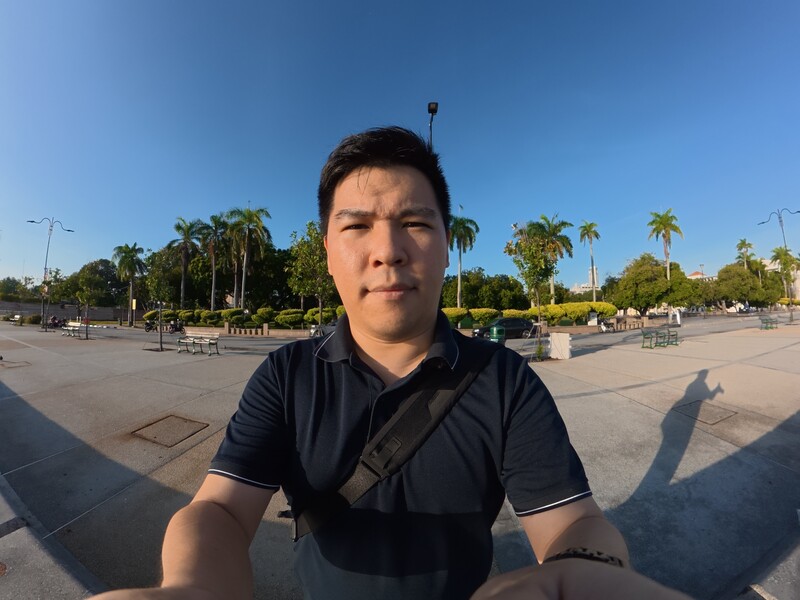 Man in dark polo shirt taking a selfie in a paved public plaza with palm trees and clear sky.
