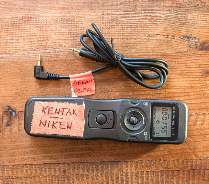 Vintage film camera with handwritten labels on wooden surface, alongside coiled power cable.