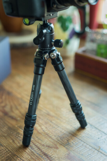 Black tripod with ball head and leg braces extended on wooden surface indoors.