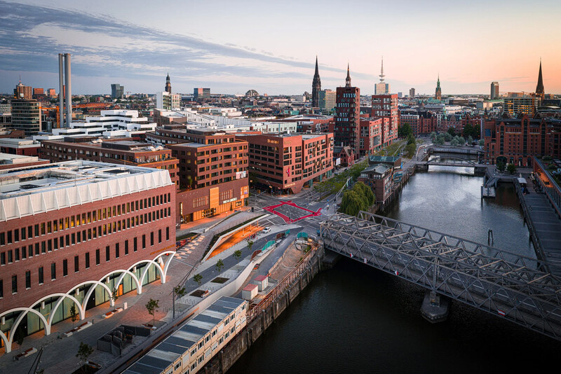 Urban waterfront cityscape at dusk with red brick buildings, church spires, and a modern pedestrian bridge over a canal.