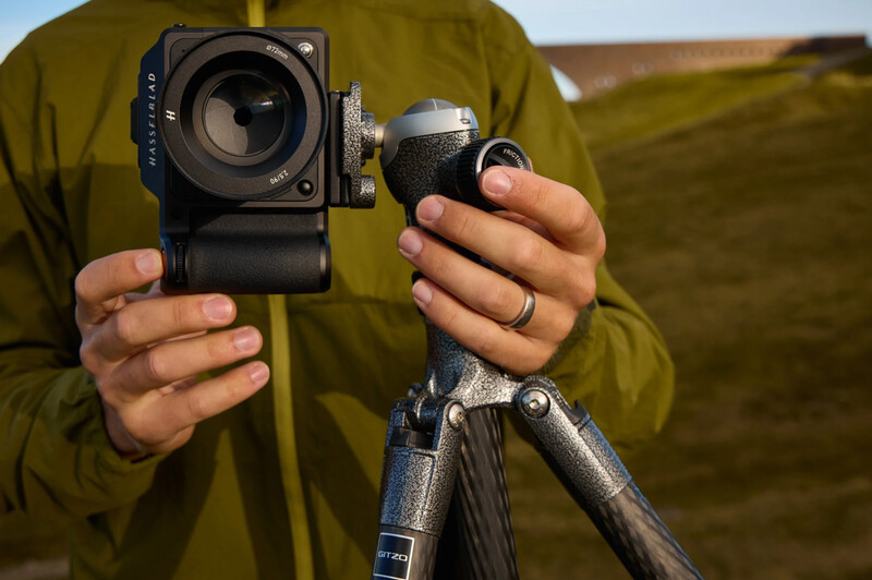Photographer in olive jacket holding a medium format camera and tripod ball head against an outdoor landscape.