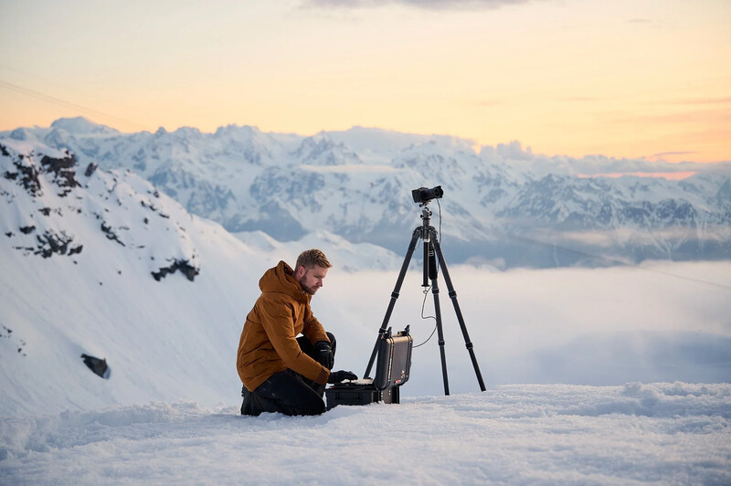 Photographer in mustard jacket operating camera on tripod atop snowy mountain peak at sunrise.