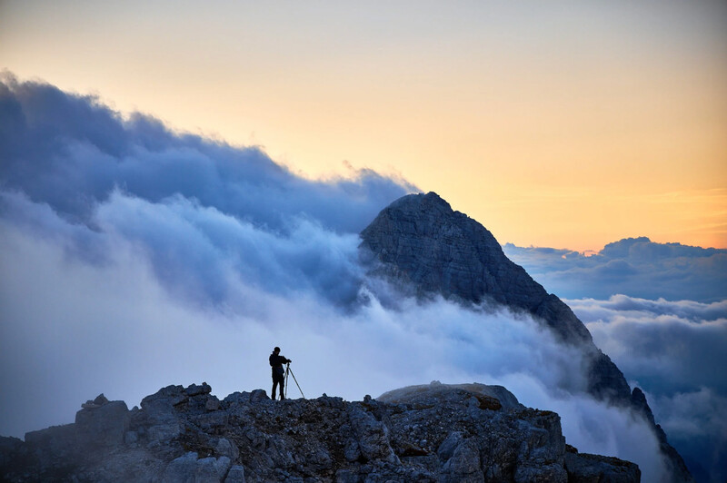 Silhouetted hiker standing on a rocky outcrop above a sea of clouds at sunrise.