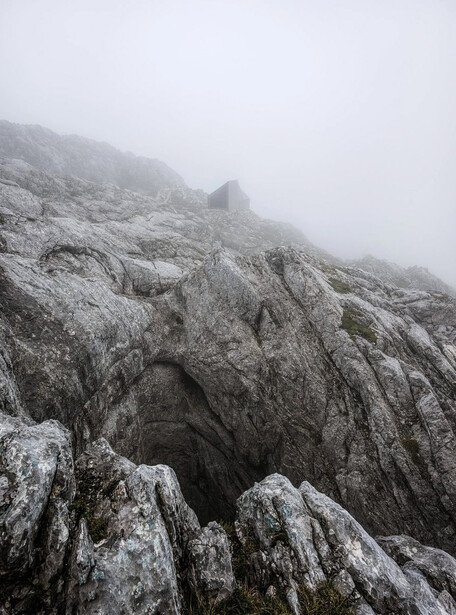 Rocky mountain ridge shrouded in dense fog and mist, with layered stone formations visible in monochrome light.