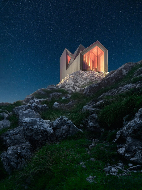Modern chapel with glowing orange interior perched on rocky hillside under starry night sky.