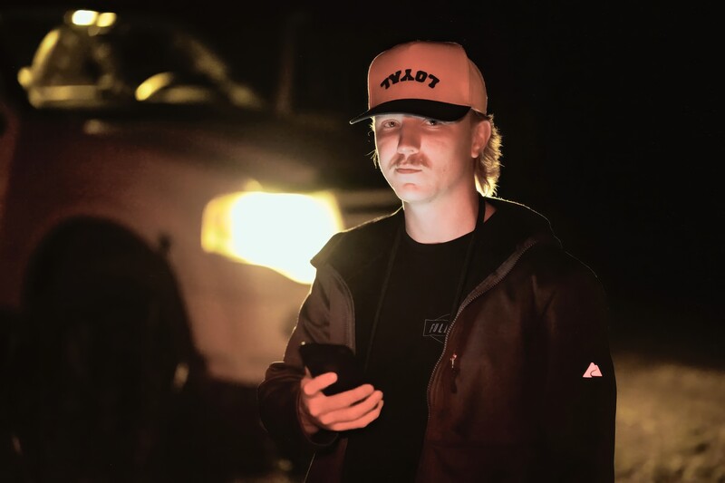 Person wearing cap and dark jacket holding phone at night with vehicle headlights in background.