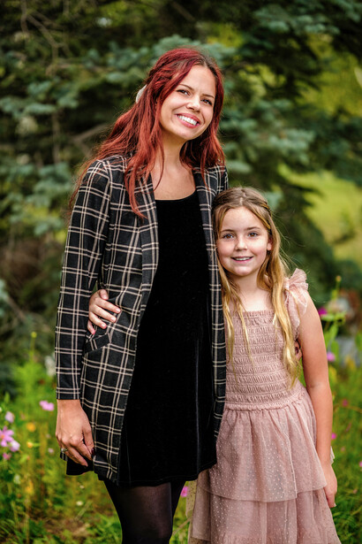 Woman with red hair and young girl standing together outdoors against green foliage background.