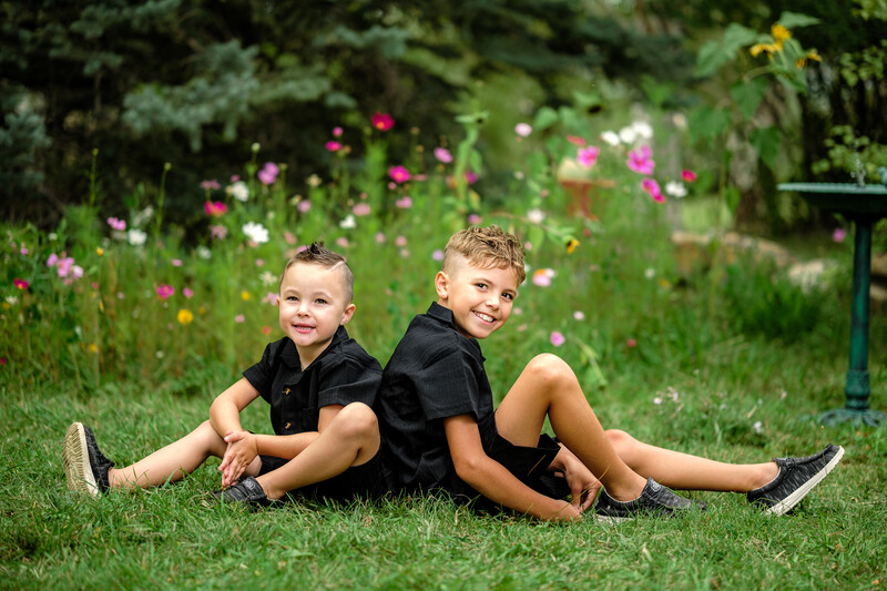 Two young boys in black shirts sitting back-to-back on grass with blooming flowers in the background.