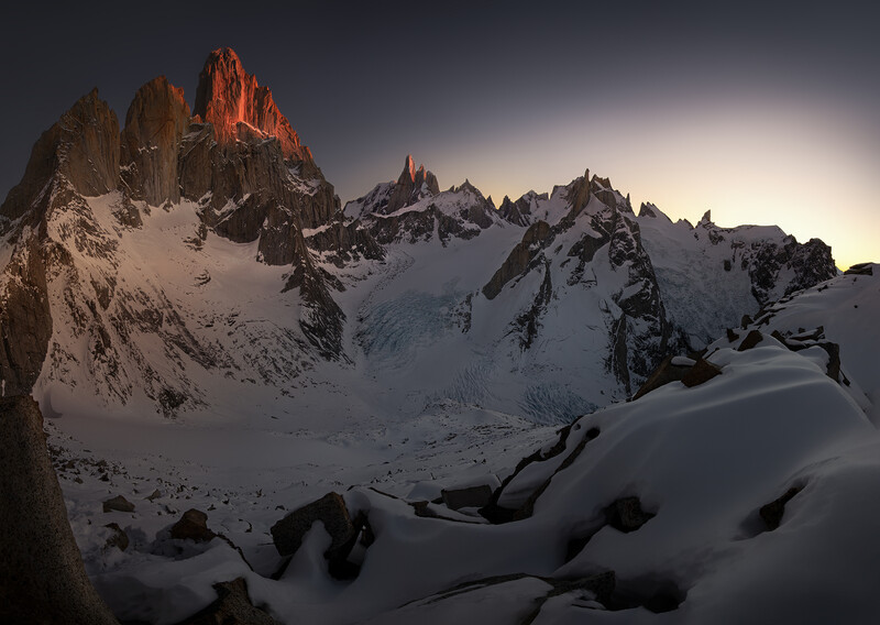 Snow-covered alpine peaks at sunset, with golden light illuminating jagged rock formations against a dark sky.