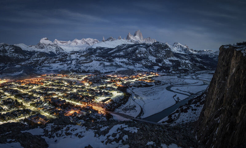 Snowy mountain peaks towering above a glowing valley town at twilight.