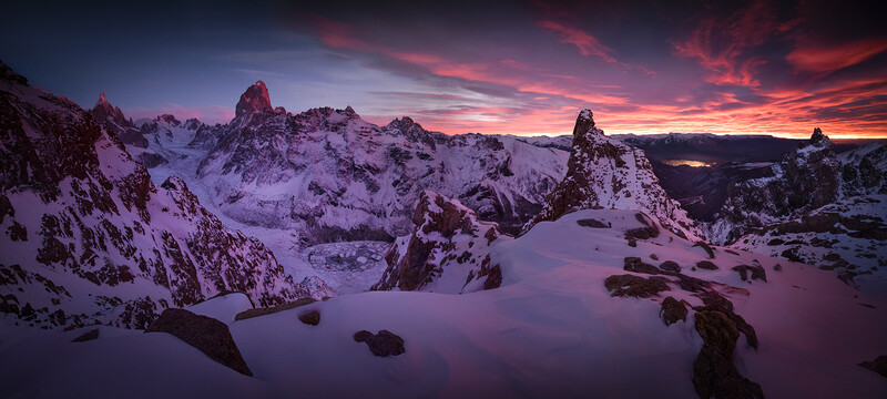 Snow-covered alpine peaks and rocky outcrops beneath a dramatic purple and orange sunset sky.